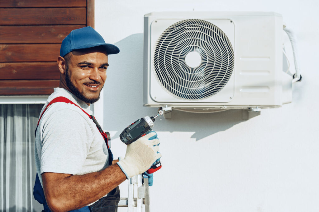 A man in overalls and a hat holds a tool beside a white air conditioner.